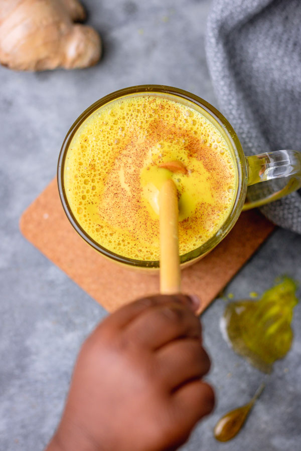 a hand holding a honey dipper in a glass of turmeric milk.