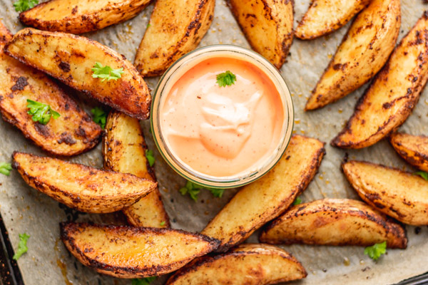 oven baked potato wedges in a lined baking sheet, garnished with chopped parsley and a dipper of spicy mayo.