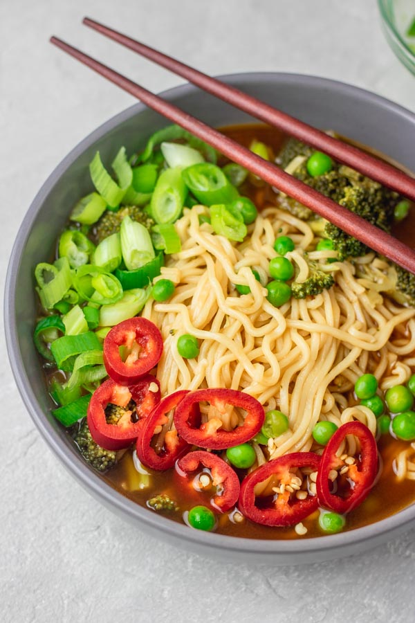 a grey bowl with ramen noodles garnished with chopped chillies and green onions. there are two chopsticks placed on the bowl.