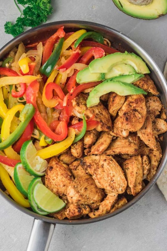 a skillet full of chicken breast pieces and sauteed peppers and onions with avocado slices.