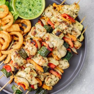 a platter of grilled chicken kabobs with curly chips, green dip and lettuce leaves.