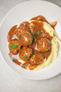 a white plate of meatballs and gravy served over mashed potato.