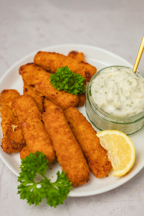 a plate of breaded fish fingers with a small pot of sauce and lemon wedge.