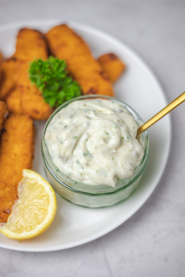 a pot of tartare sauce on a plate with fish sticks and a lemon wedge.