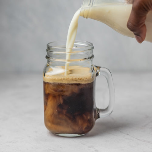 A hand pouring milk into a glass of coffee.