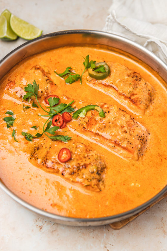 four salmon fillets in a pan of creamy coconut curry garnished with fresh chillies and parsley.