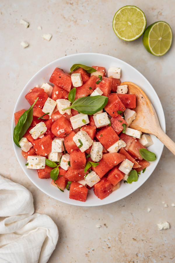 the overhead shot of a plate of watermelon salad and feta placed beside a white napkin and two lime halves.