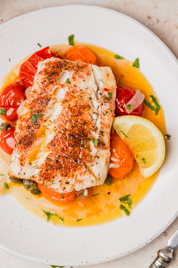 baked cod placed on juicy cherry tomatoesserved with a lemon wedge.