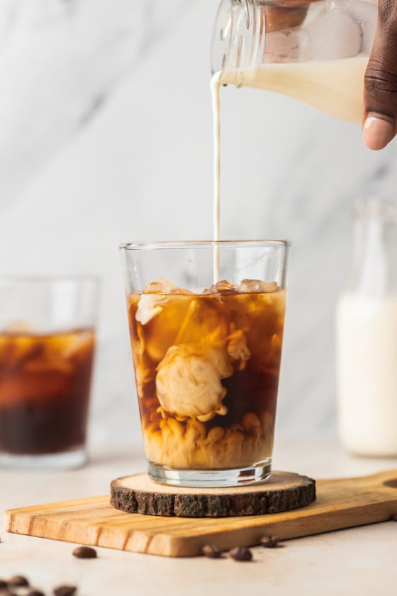 A HAND POURING MILK OVER ICED COFFEE.