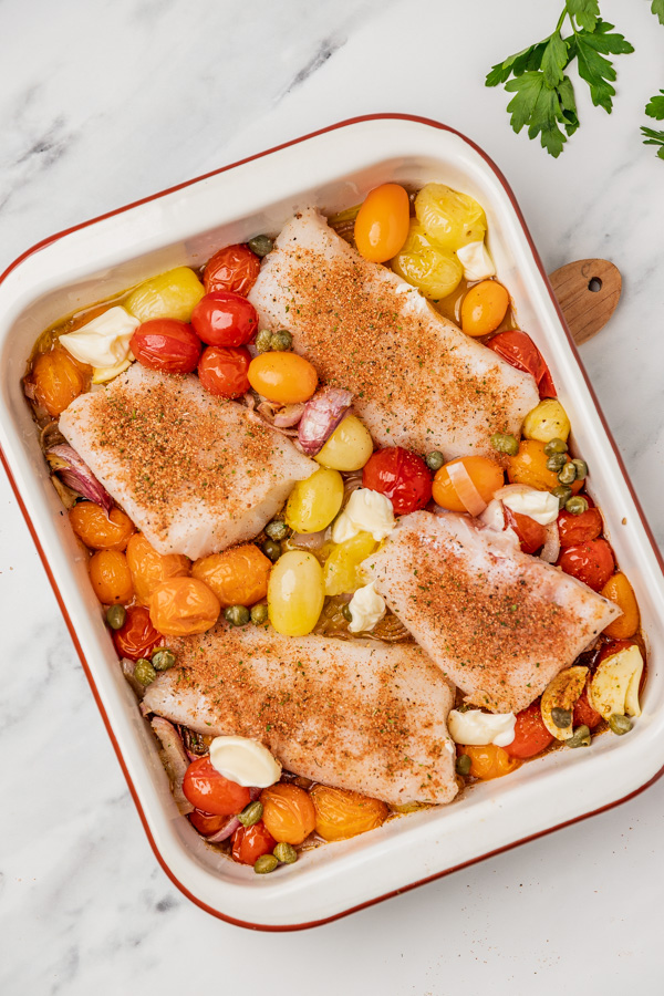 four cod fillets generously seasoned with fish seasoning placed on a bed of tomatoes in a baking tray.