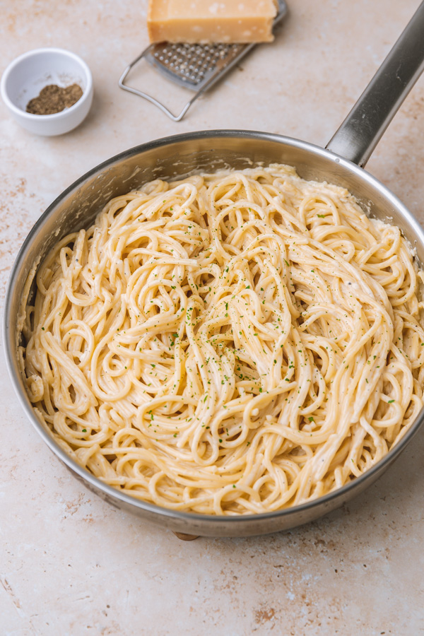 creamy spaghetti in a skillet garnished with green herbs. It is placed beside a small pot of black pepper and a block of cheese with grater.