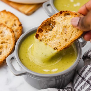 a hand dipping toasted bread into green soup.