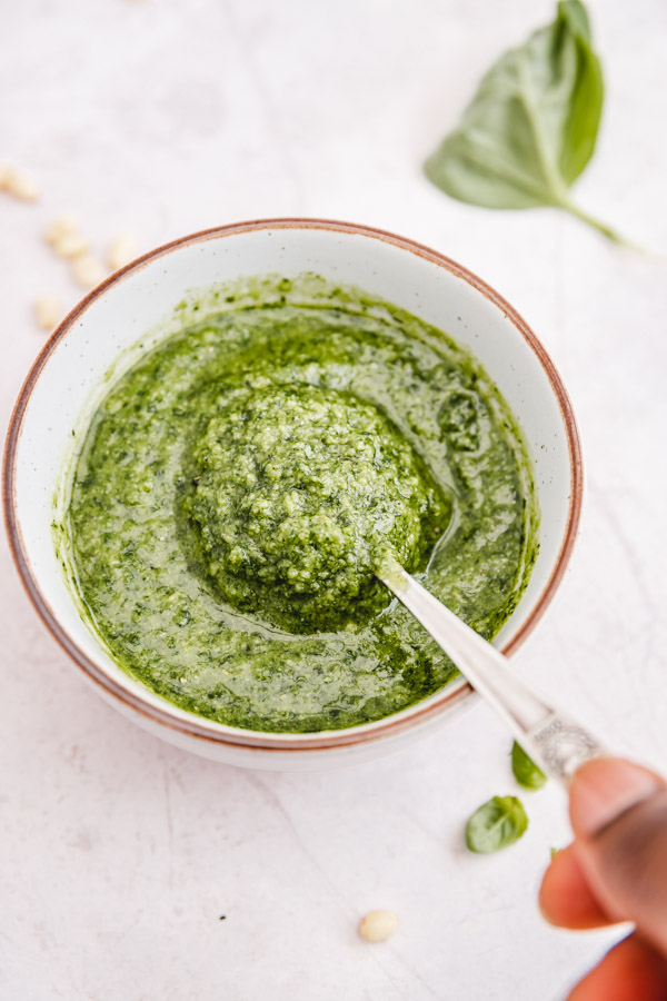 a hand about to hold up a spoon of basil pesto from the bowl.