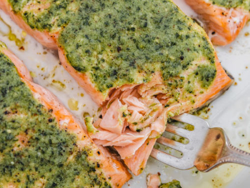freshly baked pesto salmon fillets in a baking sheet with a fork flaking the salmon fillet in the middle.