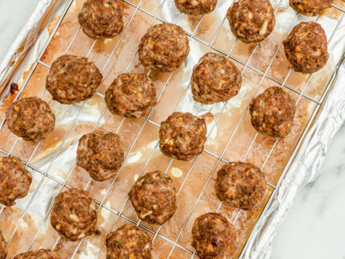 freshly baked meatballs on a wire rack placed over a lined baking sheet, the beef dripping is sitting in a the baking sheet.