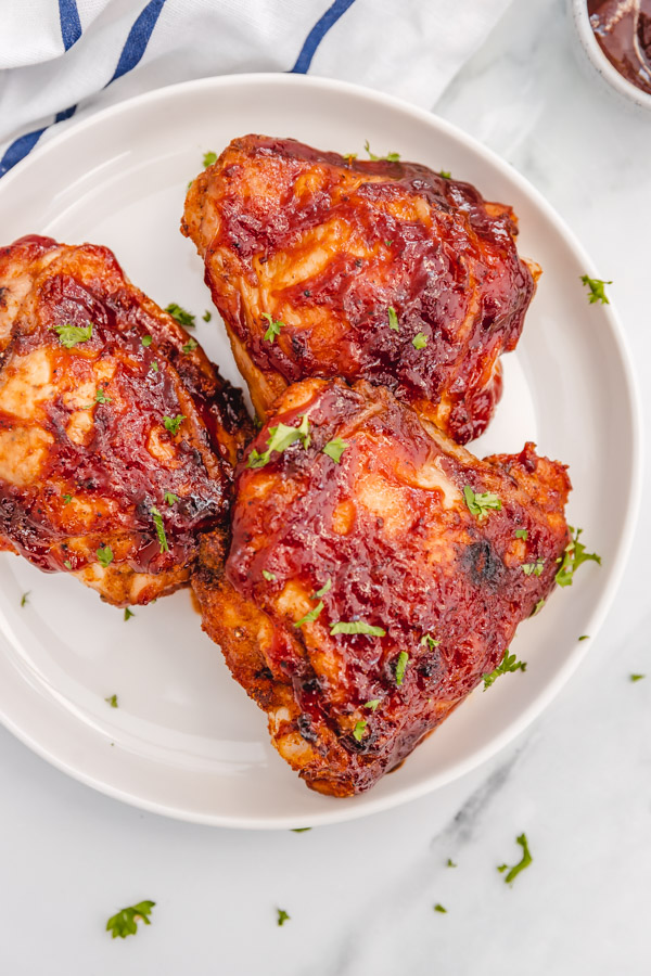 three baked bbq chicken thighs on a white plate place on a marble surface.