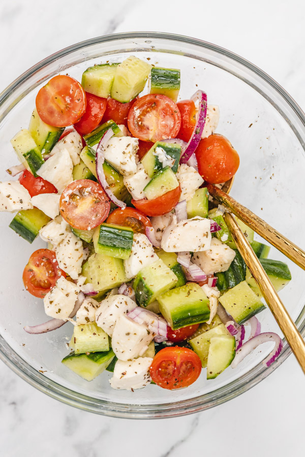 cucumber tomato mozzarella salad mixed together in a glass bowl two golden cutleries.