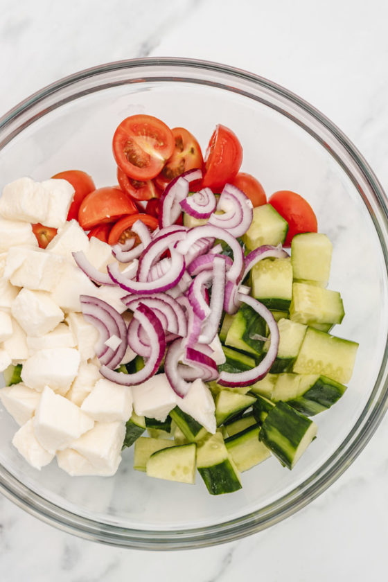 diced cucumber, oinions, chopped cherry tomatoes and diced mozzarella cheese in a large glass mixing bowl placed on  white surface.