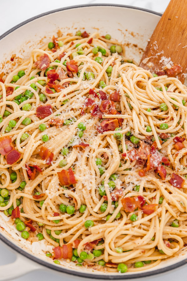 a flat wooden ladle pusing into a skillet of bacon and peas spagetti in a skillet.