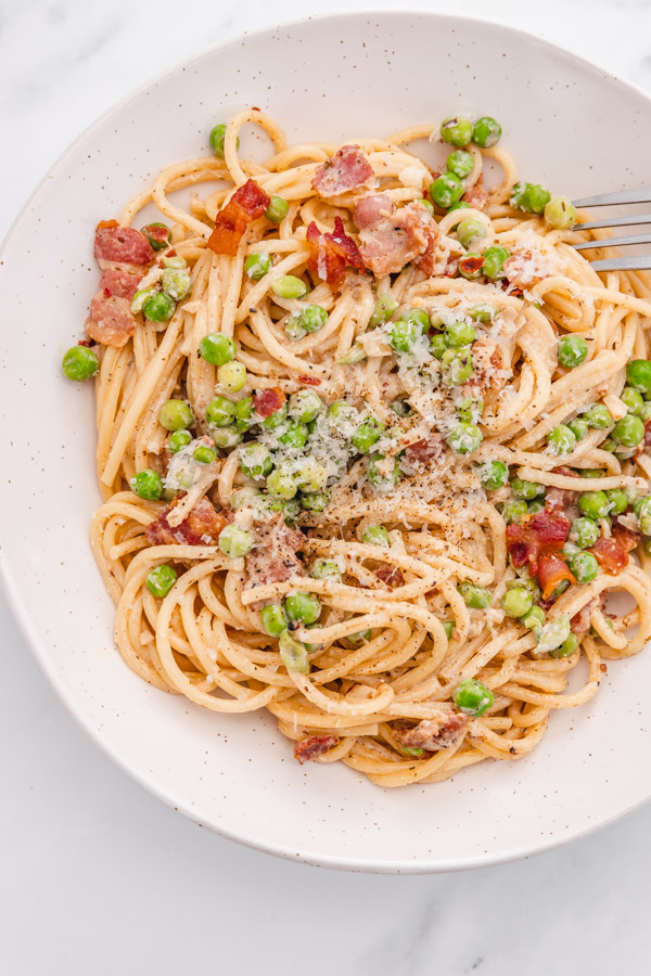 a bowl of bacon pea pasta garnished with grated parmesan cheese with a fork placed on the side of the bowl.