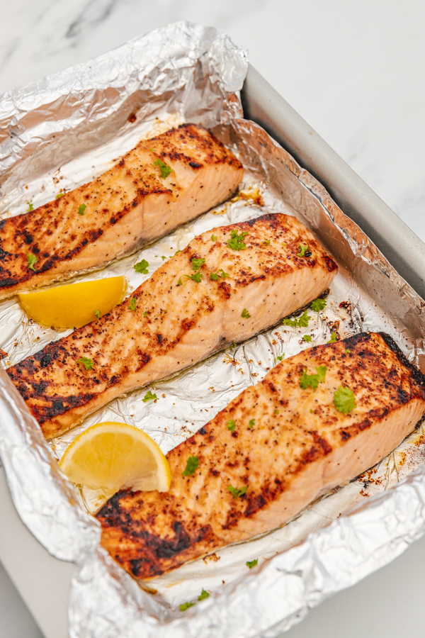 three salmon fillets in a baking sheet with two lemon wedges.