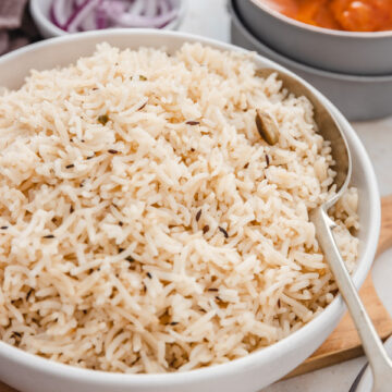 a bowl of jeera rice placed on a wooden platter placed beside a bowl or curry and sliced onions.