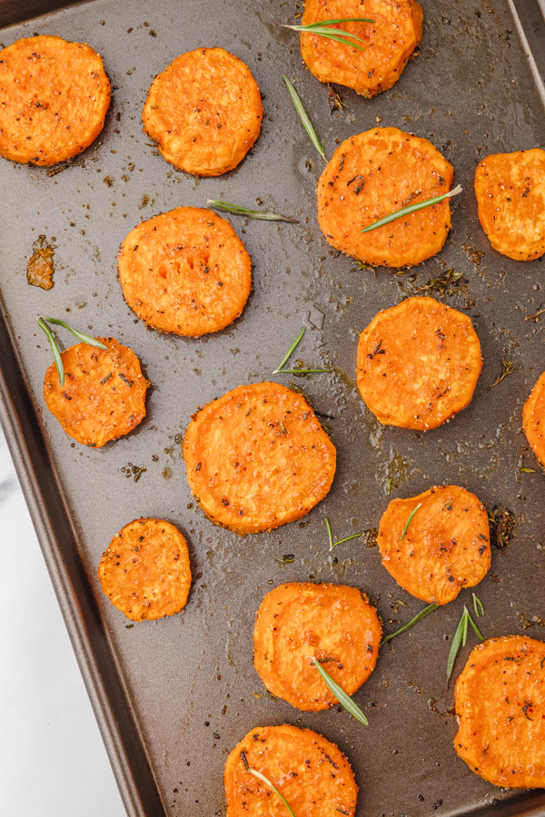 baked sweet potato clices on a baking sheet garnished with fresh rosemary leaves.