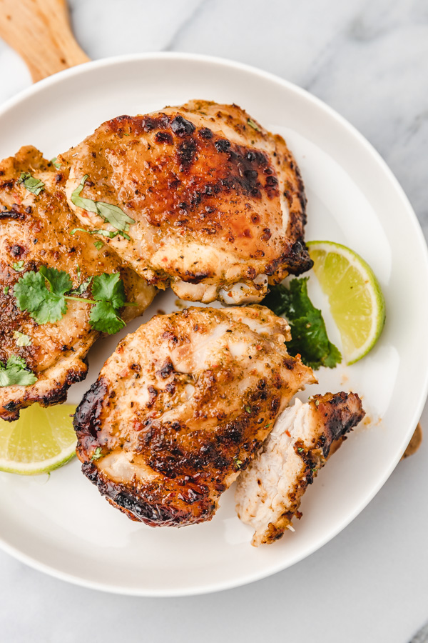 three cooked chicken thighs on a white plate with lime wedges and cilantro leaves.