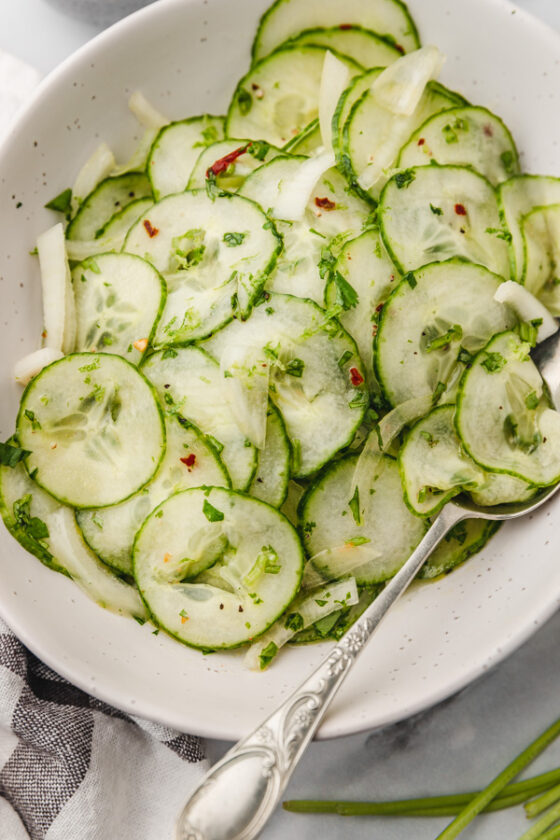 a close up of cucumber salad in a white speckled bowl with a serving spoon.