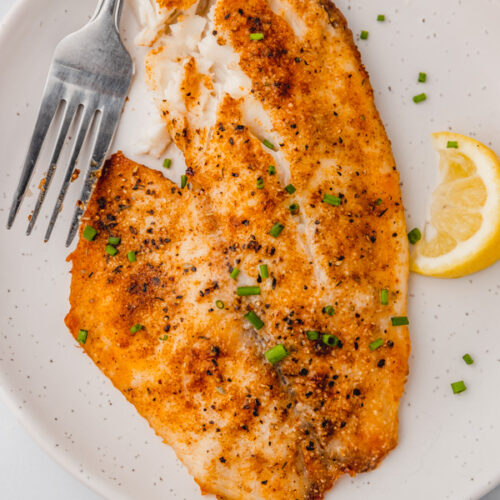 a large cooked tilapia fillet on a plate with a lemon wedge.