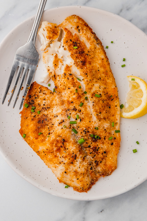 a large cooked tilapia fillet on a plate with a lemon wedge.