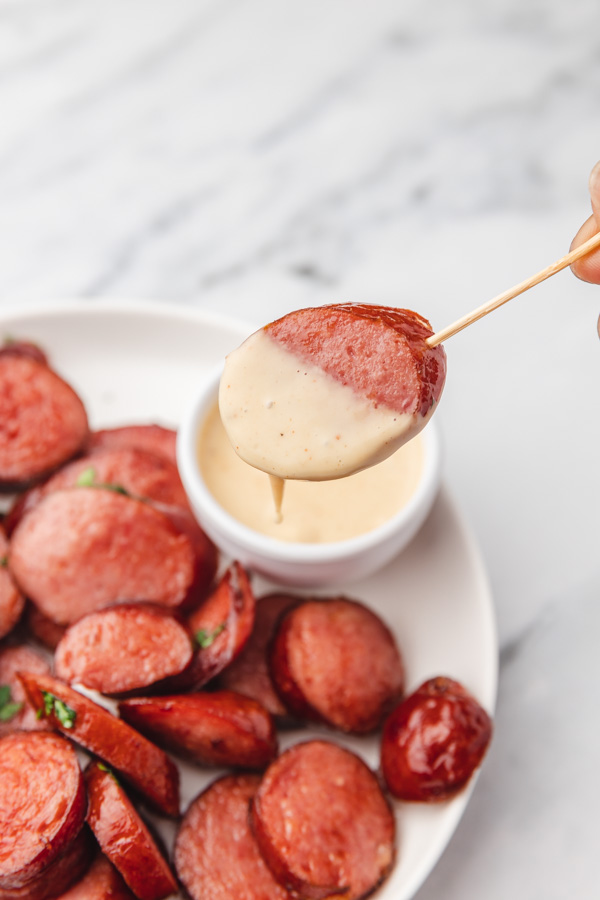 a hand holding  sliced kielbasa dipped in a sauce on a toothpick. 