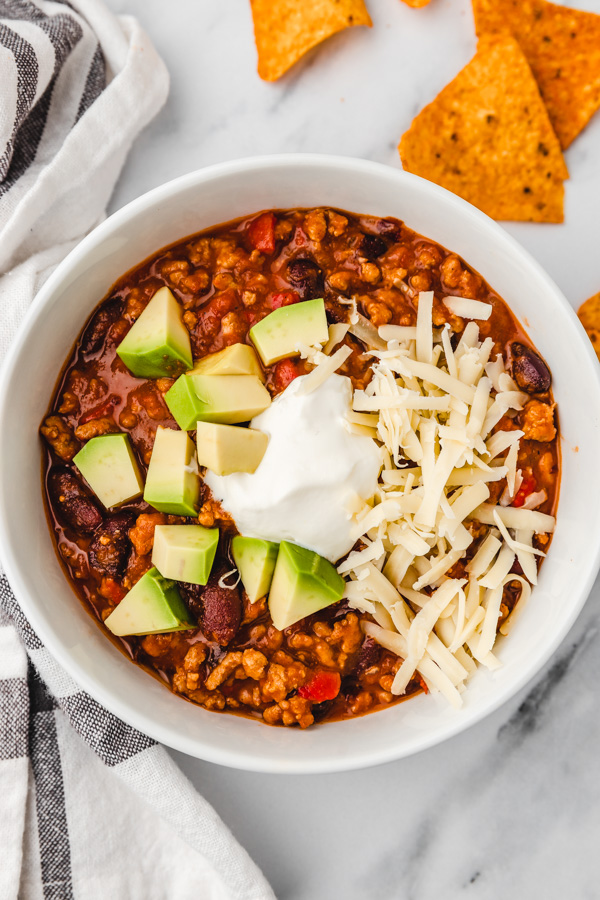 a bowl of chili con carne topped with sliced avocado, sour cream and shredded cheeese.