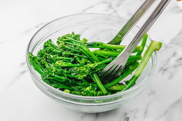 tong mixing broccolini in a bowl.