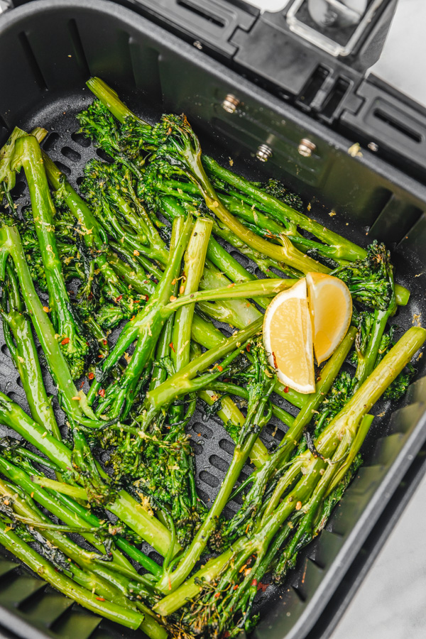 air fried broccolini in air fryer basket with two lemon wedges and red chili flakes.