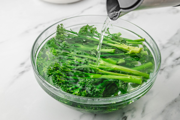 a hand pouring hot water to broccolini in a bowl.