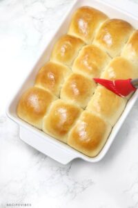 a hand brushing butter on dinner rolls in a baking dish.