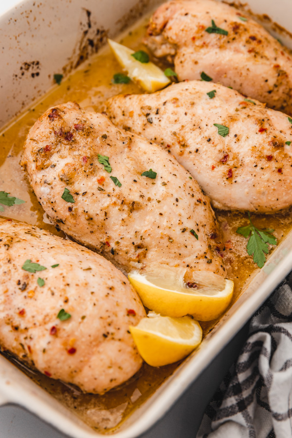four baked chicken breast in a baking dish with lemon wedges.