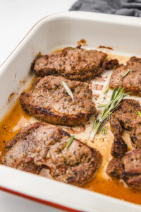 baked lamb steaks in a pan garnished with fresh rosemary leaves.