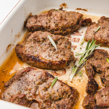 baked lamb steaks in a pan garnished with fresh rosemary leaves.
