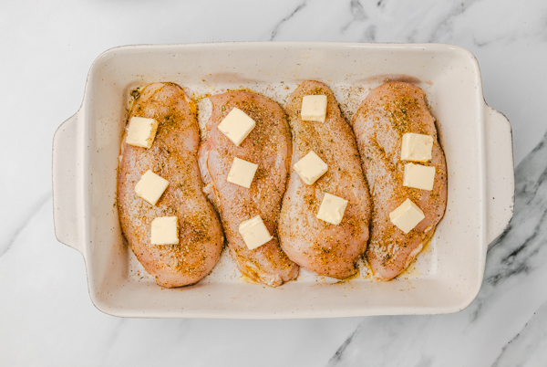 four raw chicken breasts with cubed butter in a baking dish.