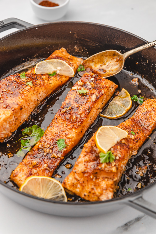 three salmon fillets sitting in a sauce in a cast iron garnished with lemon slices./ 