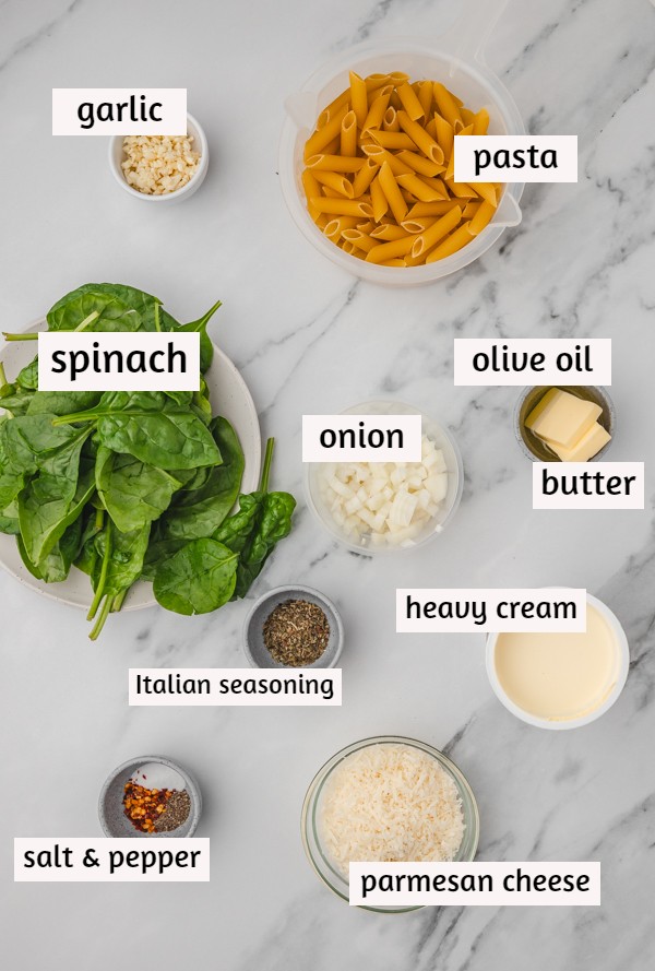 ingredients needed to make creamy spinach pasta on a white marble surface.