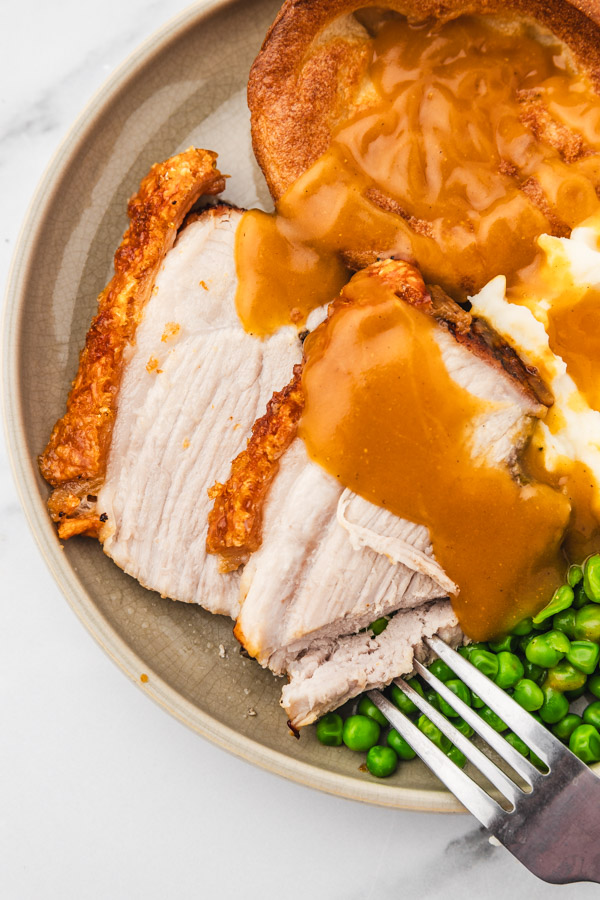 sliced pork roast with gravy and a side of peas and yorkshire pudding on a plate with a fork.