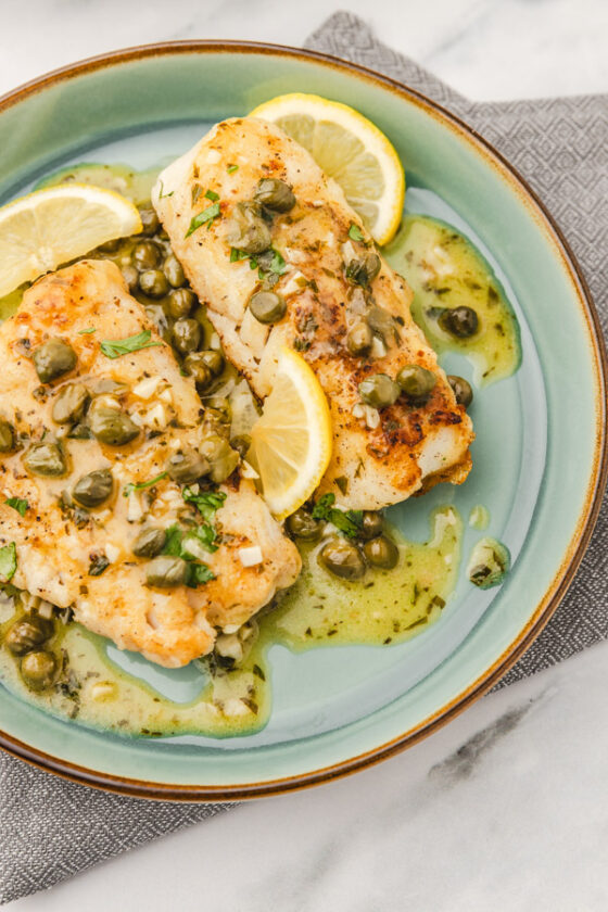two cod piccata on a plate with lemon wedges.
