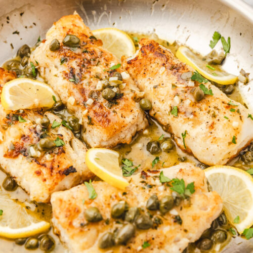 codfish piccata in a skillet garnished with fresh parsley and lemon wedges.