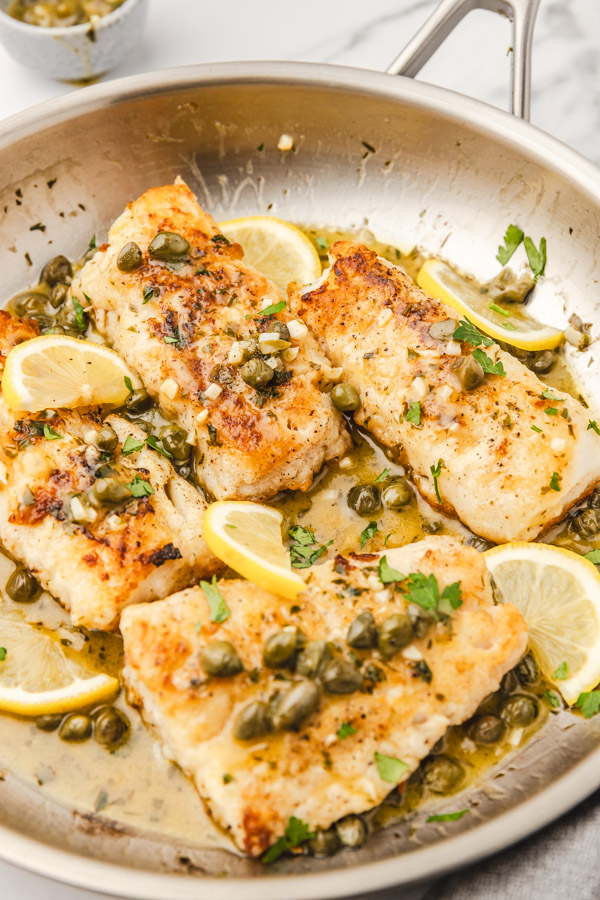 codfish piccata in a skillet garnished with fresh parsley and lemon wedges.