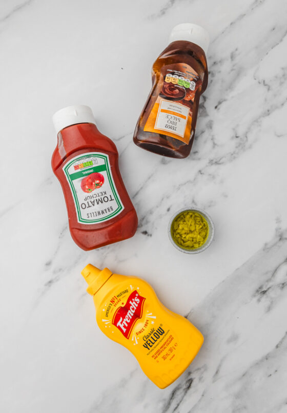 a bottle of ketchup, yellow mustard , bbq sauce and a bowl or pepperoncini on a white marble surface.
