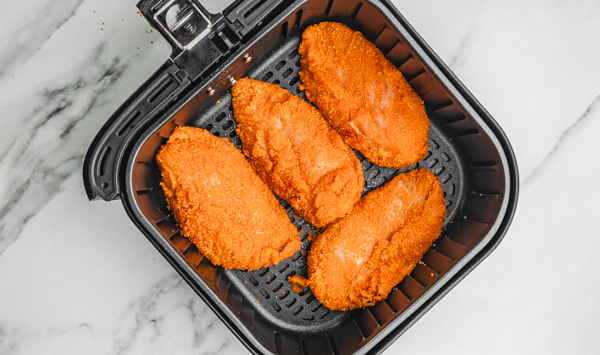 four uncooked breaded chicken breasts in an air fryer basket.