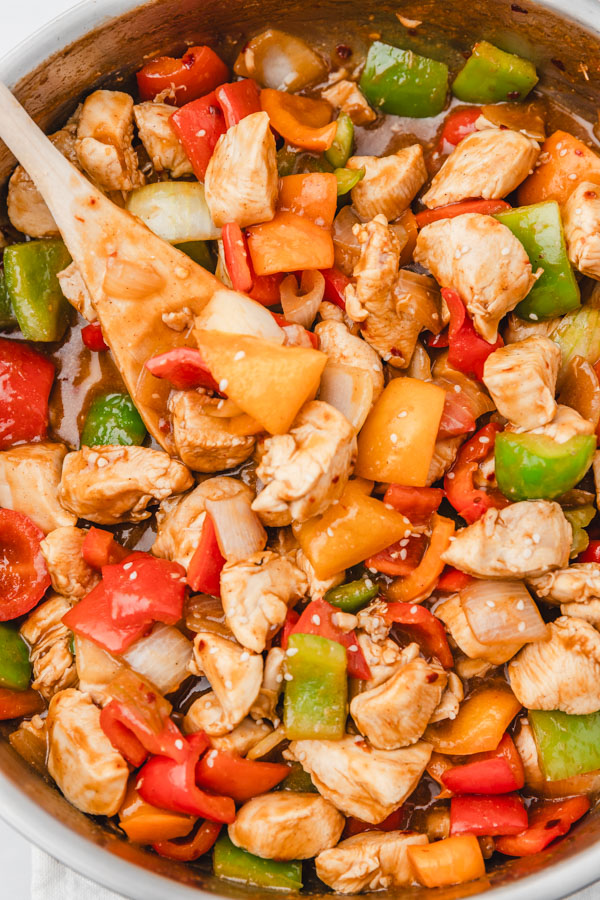 a wooden ladle pushing chicken and peppers stir fry in a skillet.